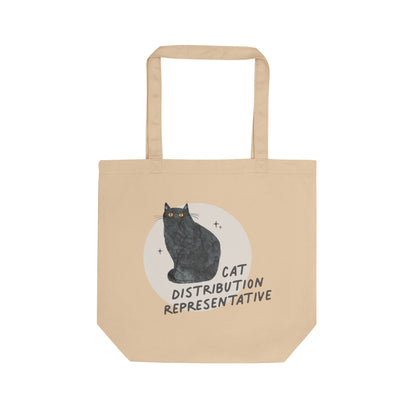 A beige canvas tote bag with two handles, featuring a graphic of a black cat sitting in front of a white circle with the text 'CAT DISTRIBUTION REPRESENTATIVE'.