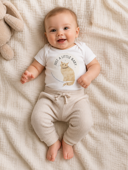 A baby lying on a textured cream muslin blanket, wearing a white short-sleeve bodysuit with a cat graphic and the text 'JUST A LITTLE BABY' paired with ribbed cream pants.