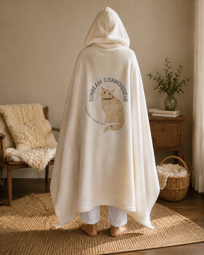 A person stands with their back to the camera wearing a cream-colored hooded blanket or robe with a cat graphic and the text 'SUNGEARI CONNOISSEUR' printed on the back, in a cozy, sunlit room.