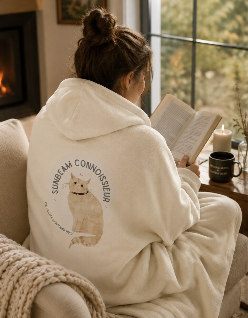 A woman sits on a couch in a cozy living room, reading a book while wrapped in a cream fleece hoodie. The hoodie features a large graphic on the back with a cat illustration and the text 'SUNBEAM CONNOISSEUR'.