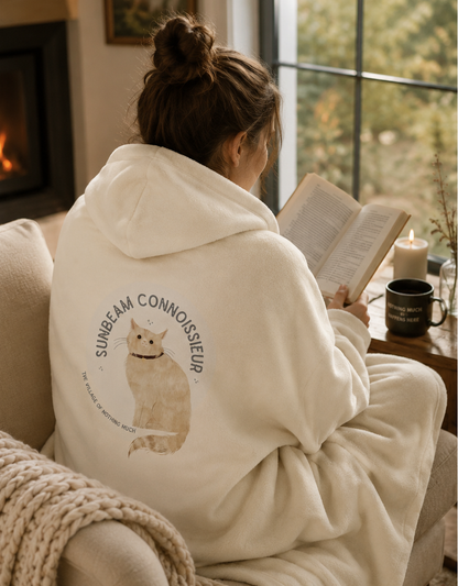 A woman sits on a couch in a cozy living room, reading a book while wrapped in a cream fleece hoodie. The hoodie features a large graphic on the back with a cat illustration and the text 'SUNBEAM CONNOISSEUR'.