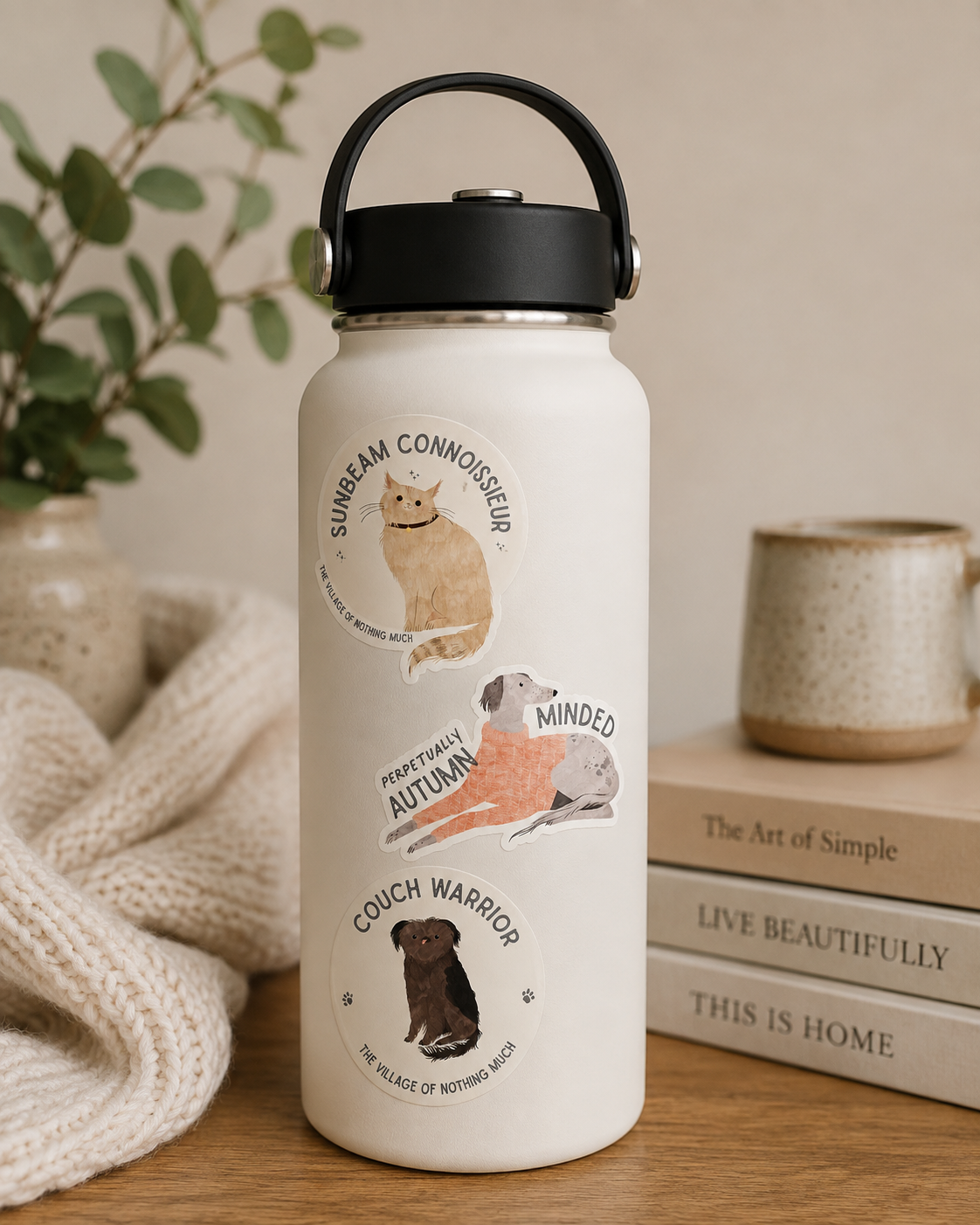 A cream-colored insulated water bottle with a black lid and handle, decorated with three circular dog-themed illustrations and text, set on a wooden table next to a knitted blanket, books, and a ceramic mug.