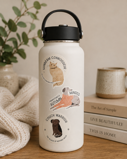 A cream-colored insulated water bottle with a black lid and handle, decorated with three circular dog-themed illustrations and text, set on a wooden table next to a knitted blanket, books, and a ceramic mug.
